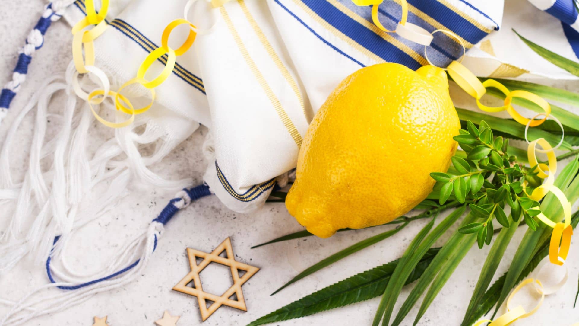 A yellow etrog fruit, lulav branches, a tallit with blue and gold stripes, yellow paper chains, and a wooden Star of David on a light surface, celebrating the Jewish festival of Sukkot. Shalom Y’all!.