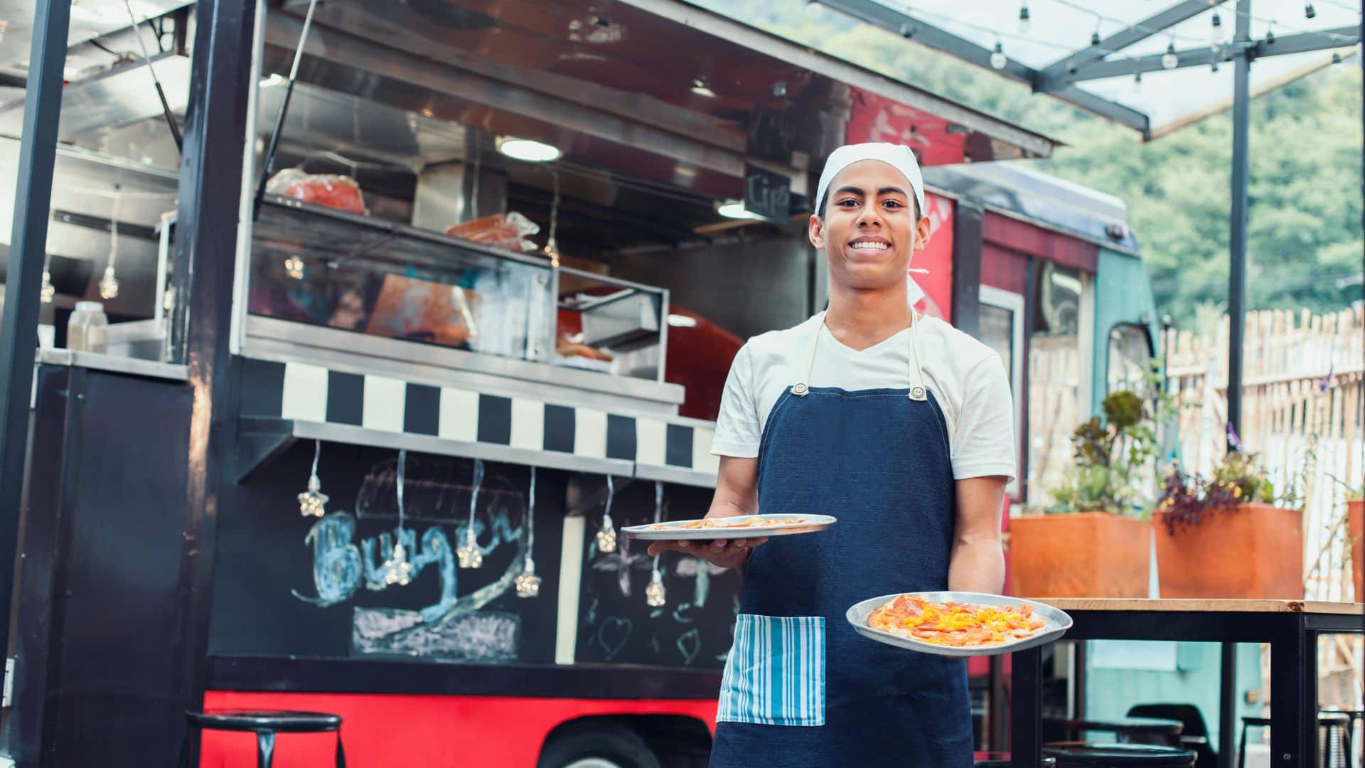 A smiling food truck worker in a white shirt, navy apron, and chef hat holds two plates of pizza outside a colorful Food Truck at Eastern Wharf with a black-and-white striped counter.