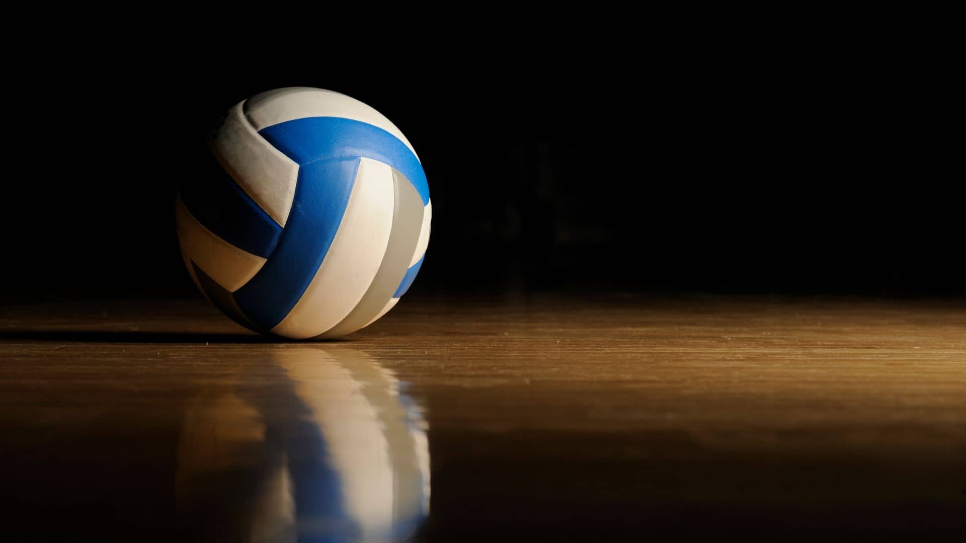 A blue and white volleyball rests on a polished wooden floor, its dramatic shadow and reflection captured against a dark background—echoing the intensity of SEC Women's Volleyball competition.