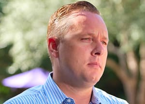 A man with short, light brown hair wearing a blue and white striped shirt stands outdoors with a neutral expression. Trees and blurred objects are in the background.