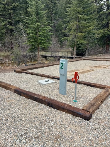 A gravel campsite with wooden borders features a water spigot, electrical hookup labeled "2," and a metal cover, surrounded by trees and a small footbridge in the background.