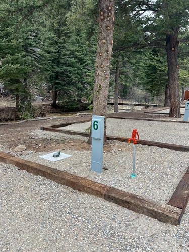 An empty gravel campsite with a utility hook-up labeled 6, a water spigot, and a large tree nearby, surrounded by pine trees in a forested area.