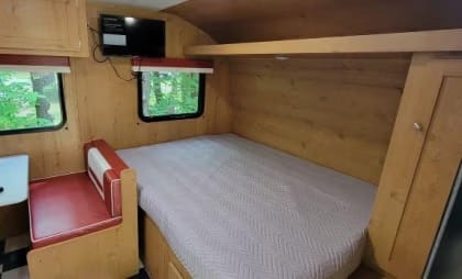 Compact camper interior with bed, seating area, and wood paneling.