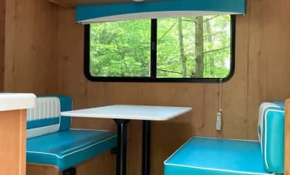 Interior of a camper with a dining area, featuring a table and cushioned benches, and a window overlooking a wooded area.
