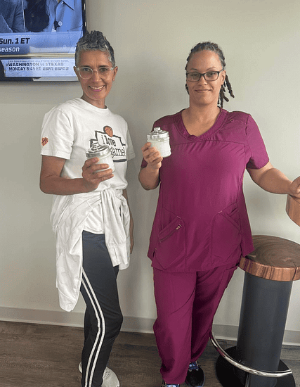 Two women stand indoors; one wears a white T-shirt and track pants, the other wears purple scrubs. Both are holding glass jars and smiling at the camera.