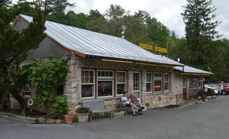 Image of a rustic brick building with a metal roof housing a restaurant. Two people sit on a bench near the entrance, surrounded by greenery and parked cars. Trees are visible in the background, enhancing the Blue Ridge Beauty of this charming location.