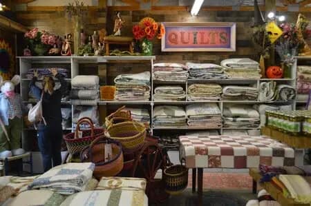 A person browsing quilts in a store filled with shelves of diverse patterns, quaint baskets, and a colorful "QUILTS" sign on the wall. One quilt features stunning Shenandoah Views while another captures the essence of Blue Ridge Beauty.