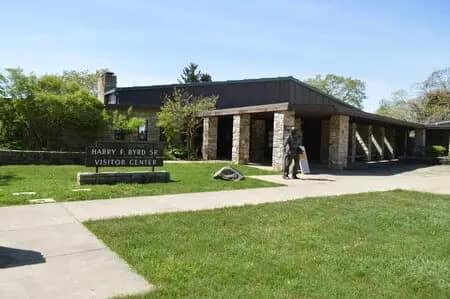 The image shows the Harry F. Byrd Sr. Visitor Center, with a stone exterior and a pathway in front. A person walks towards the entrance on a sunny day, surrounded by the Blue Ridge Beauty and under Spacious Skies Shenandoah Views.