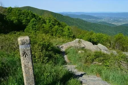 A stone marker with the Appalachian Trail logo stands beside a narrow dirt path, offering captivating Shenandoah Views with lush green hills and distant mountains in the background.