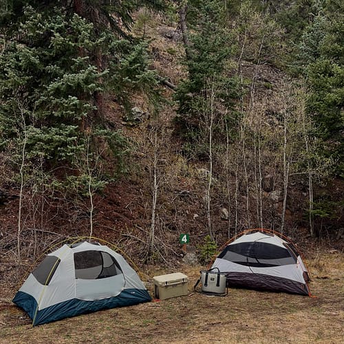 Two camping tents are set up on grass near a forested hillside, with two coolers placed between them. A green sign with the number 4 is visible behind the tents.