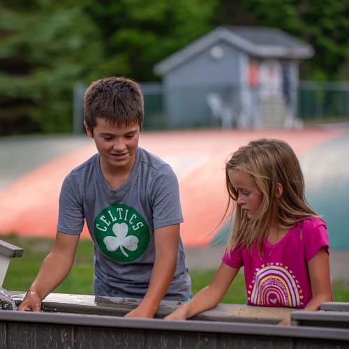 Two children are outdoors, standing beside each other at a water play area. The boy, wearing a grey "Celtics" t-shirt, is focused on an activity. The girl beside him, in a pink shirt with a rainbow design, is also engaged. Green trees and a small building are in the background.