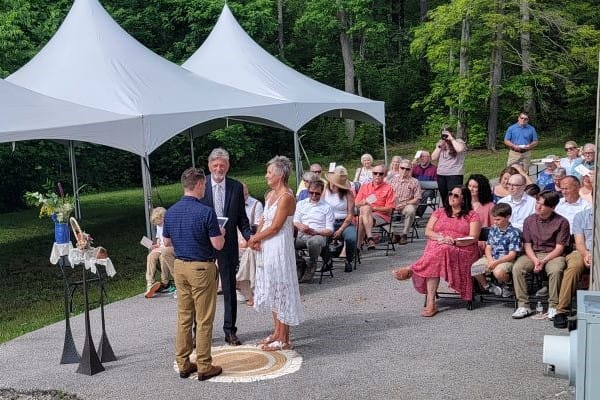 A couple stands hand in hand at a picturesque outdoor wedding ceremony. The officiant reads from a book, as guests seated under two white tents in the lush forested area attentively watch the event unfold. The scene is bright and filled with greenery, capturing the beauty of weddings perfectly.