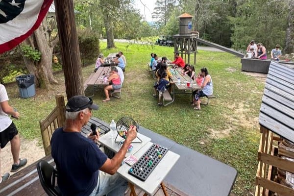 A group of people sits at picnic tables playing bingo outdoors, enjoying a leisurely event. A man is calling numbers from a small stage. A playground slide and trees in the background enhance the relaxed, park-like setting, perfect for casual gatherings or even informal weddings.