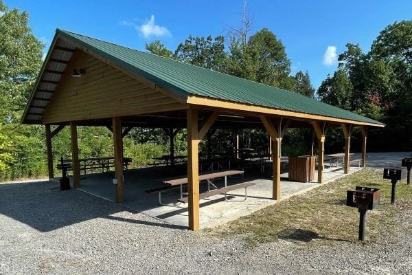 A wooden pavilion with a green roof in a park setting, perfect for events or weddings, surrounded by trees. Picnic tables sit underneath, and nearby charcoal grills are ready on a gravel surface. The sky is clear and sunny, completing this idyllic scene.