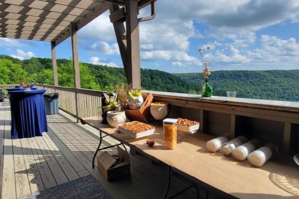 A wooden deck overlooking a lush green valley and a clear blue sky is beautifully set for events. The tables are adorned with snacks, glass jars, and decorations, perfect for intimate weddings. In the background, a tall table with a blue tablecloth adds elegance to the enchanting setup.