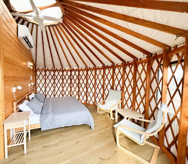 Image shows the interior of a cozy yurt with a wooden lattice structure and a light wood floor. The space contains a neatly made bed, a small bedside table with a lamp, and two white cushioned lounge chairs. A ceiling fan and air conditioner are installed for comfort.