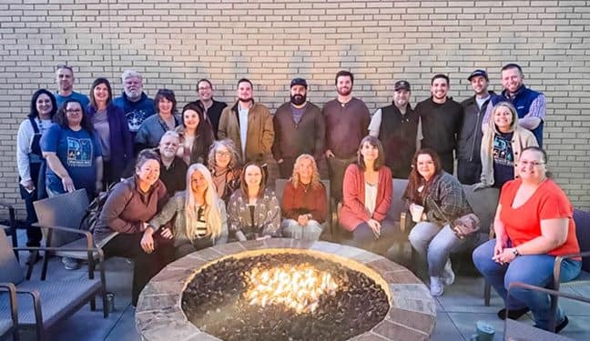 A group of people are gathered around an outdoor fire pit at Spacious Skies Campgrounds. They are smiling and posing for the camera. The background is a brick wall, and there are chairs surrounding the fire pit, capturing the relaxed vibe of their N.C. staff retreat.