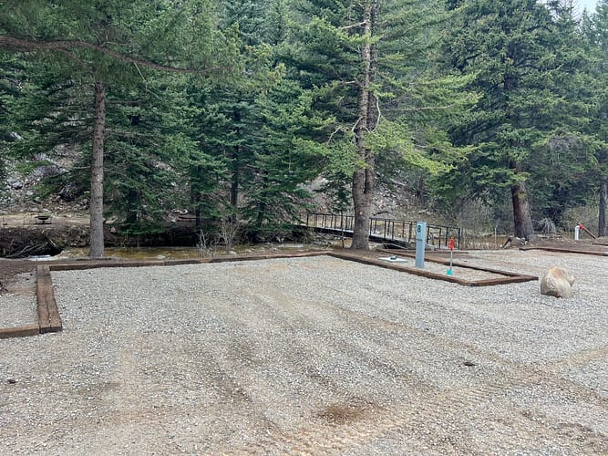 Gravel campsite with utility hookups surrounded by tall pine trees. A small wooden footbridge crosses a creek in the background, and a large rock sits near the edge of the site.