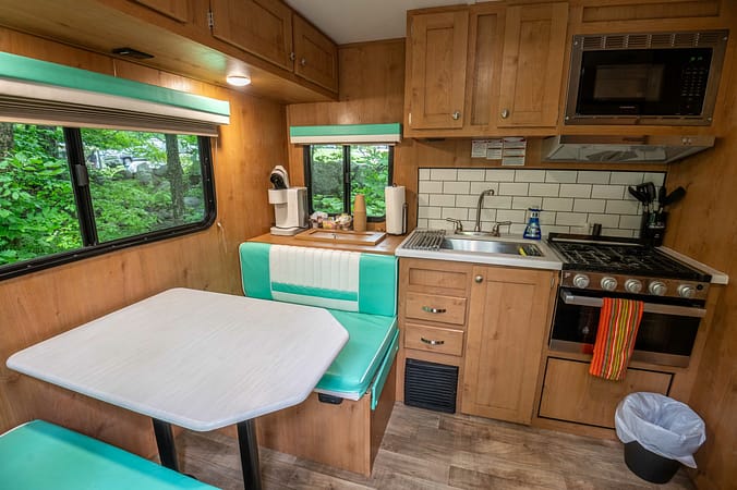 A cozy RV interior featuring a compact kitchen with wooden cabinets, a microwave, stove, sink, and counter space. Adjacent is a dining area with a light blue cushioned bench and table beside a window. The decor includes a tiled backsplash and modern appliances.
