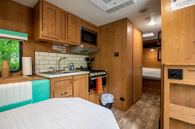 Interior of a cozy camper featuring a compact kitchen with wooden cabinets, a stove, a microwave, and a small sink. A dining table with seating is in the foreground. A narrow hallway leads to a sleeping area with a bed. The decor has a rustic wooden theme.