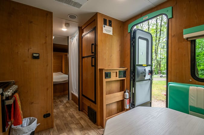 Interior of a camper with a wooden finish. Features include an open door leading outside, a bed visible through a doorway, a vertical cabinet, and a mounted fire extinguisher. The scene is well-lit by natural light entering through windows and the door.