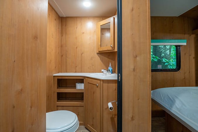 A small, wood-paneled bathroom in an RV with a toilet, a countertop holding a bottle of hand sanitizer, an upper cabinet with a mirrored door, and an open storage area beneath. A nearby window with a green exterior view and part of a bed are also visible.