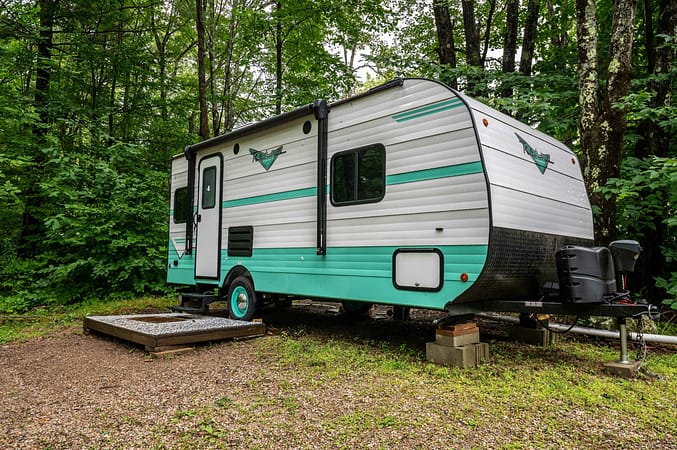 A compact, mint green and white travel trailer is parked on a gravel pad in a lush, wooded campsite. The trailer features a single entrance door, several windows, and is supported by concrete blocks and a propane tank at the hitch.