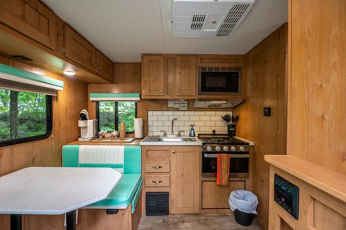 A cozy RV kitchen and dining area featuring wooden cabinets and walls. The kitchen includes a stove with an oven, a microwave, a sink, and a white tiled backsplash. The dining area has a light blue cushioned bench and table. A window offers a view of greenery.