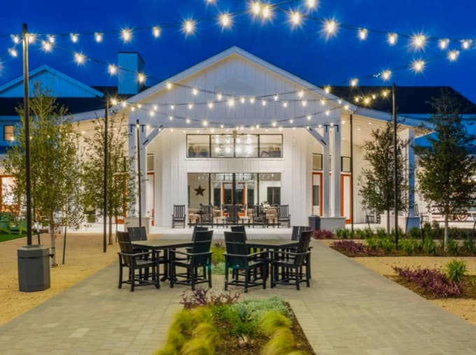 Outdoor seating area with black tables and chairs is shown in front of a white building at night, illuminated by overhead string lights and surrounded by small trees and landscaping.