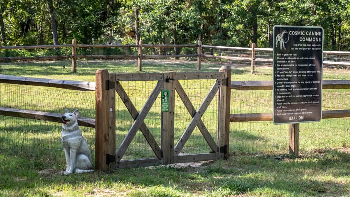 A white dog statue sits next to a wooden gate at the entrance of a fenced dog park. A sign near the gate reads "Cosmic Canine Commons," detailing park rules. The area is surrounded by green grass and trees, with another fence in the background.