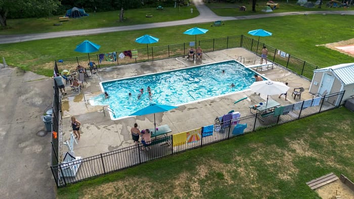 Aerial view of a rectangular outdoor swimming pool with people swimming and relaxing. The pool is surrounded by a fence and features blue umbrellas and lounge chairs. Green grass and a few trees are visible in the surrounding area.