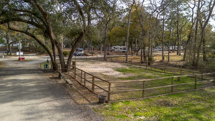 A fenced, gravel RV campsite sits among tall trees in a wooded area, with several RVs parked in the background. The ground is partly grassy, and the atmosphere appears peaceful and quiet.