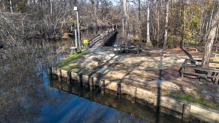 A wooden dock and picnic area sit beside a calm river, surrounded by bare trees in winter. A wooden bridge stretches over the water, and benches and tables are set up on the sandy ground.