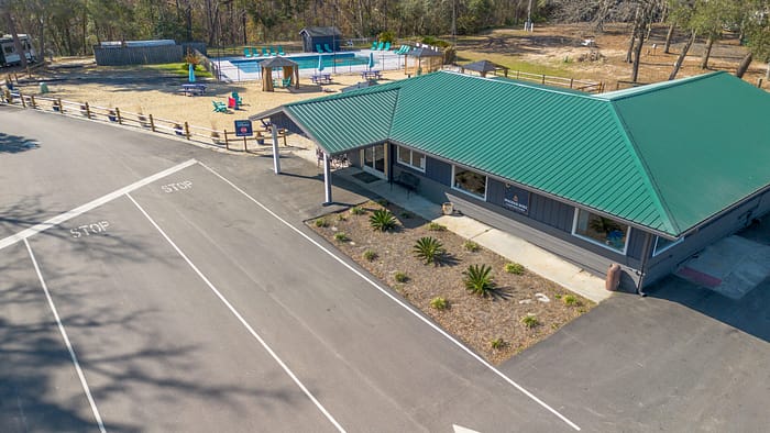 Aerial view of a building with a green roof next to a paved road with "STOP" markings, a small landscaped area, and a playground with a pool, swings, and slides in a fenced area behind the building. Trees surround the area.