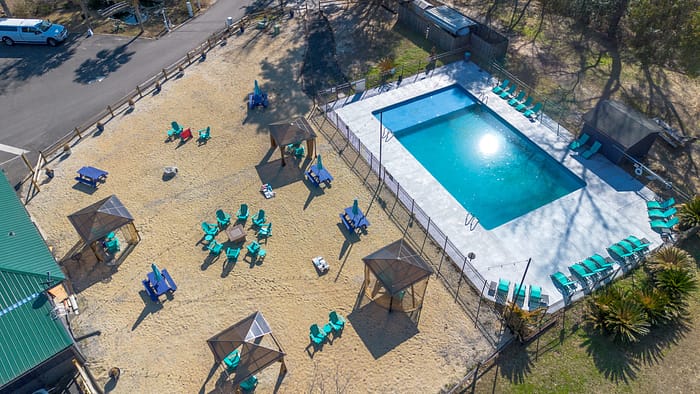 Aerial view of a fenced outdoor swimming pool surrounded by lounge chairs and shaded cabanas on sandy ground, with several seating areas and a nearby road.