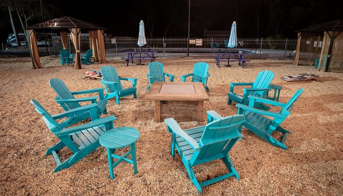 A group of turquoise Adirondack chairs arranged in a circle around a fire pit on a gravel surface, with gazebos, picnic tables, and umbrellas in the background at night.