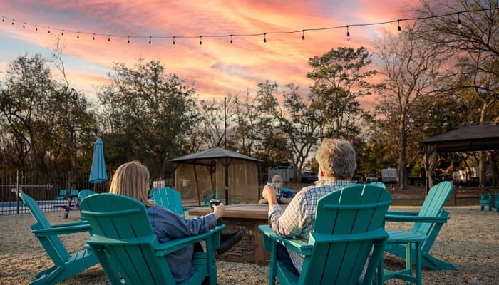 Two people sit in turquoise Adirondack chairs around a firepit, enjoying drinks at sunset. String lights hang above, and trees with sparse leaves are in the background, creating a relaxed outdoor atmosphere.