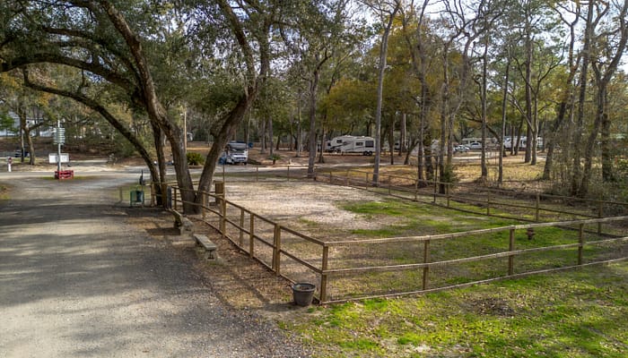 A fenced, gravel RV campsite sits among tall trees in a wooded area, with several RVs parked in the background. The ground is partly grassy, and the atmosphere appears peaceful and quiet.