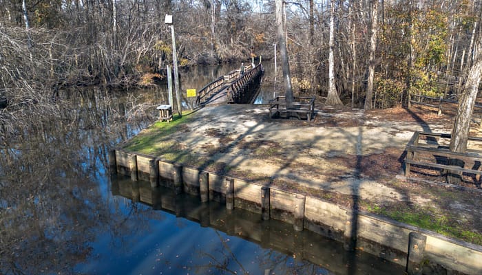 A wooden dock and picnic area sit beside a calm river, surrounded by bare trees in winter. A wooden bridge stretches over the water, and benches and tables are set up on the sandy ground.