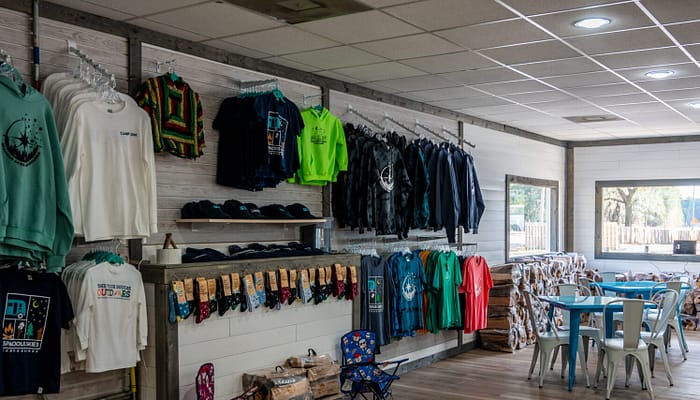 A gift shop with racks of colorful t-shirts, sweatshirts, hats, and bandanas on the left wall. On the right, there are tables and chairs by windows letting in natural light. The shop has wooden floors and a cozy atmosphere.