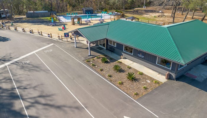 Aerial view of a building with a green roof next to a paved road with "STOP" markings, a small landscaped area, and a playground with a pool, swings, and slides in a fenced area behind the building. Trees surround the area.