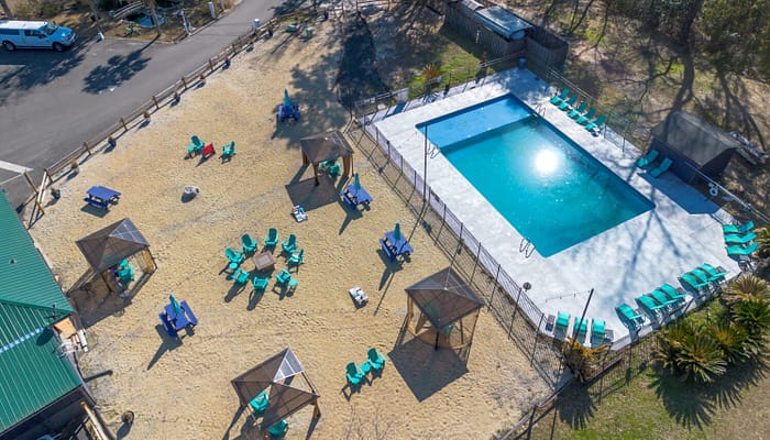 Aerial view of a fenced outdoor swimming pool surrounded by lounge chairs and shaded cabanas on sandy ground, with several seating areas and a nearby road.