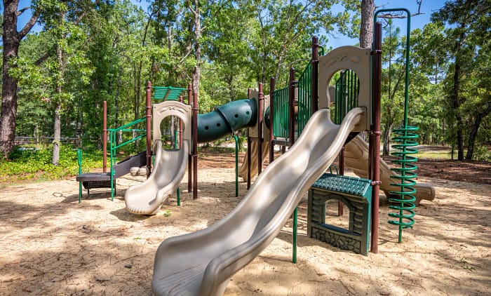 A playground set in a park with slides, climbing equipment, and a tunnel, surrounded by trees. The ground is covered in sand, and it's a sunny day.