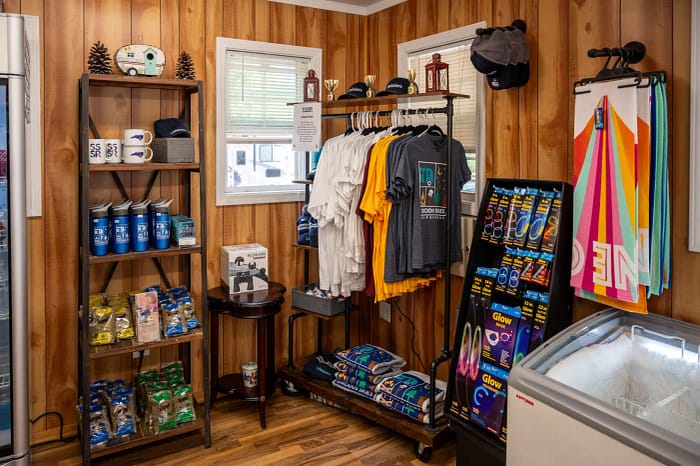 A small store with wooden walls sells various items. It has shelves displaying water bottles, mugs, hats, and snacks. Hanging racks have t-shirts. A corner stand displays flip-flops and glow sticks. A glass display case on the right is closed.