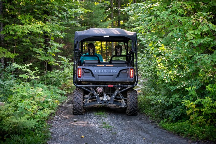 A Honda off-road vehicle with two seats, viewed from the back, driving on a gravel trail through a dense, green forest. The vehicle is surrounded by lush foliage and trees on a sunny day.