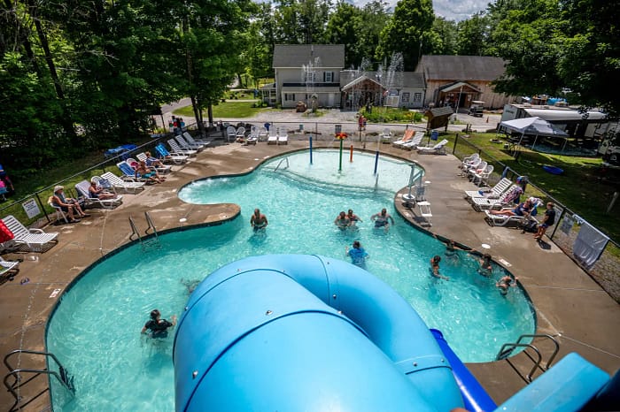 Aerial view of a swimming pool with a blue slide, surrounded by people lounging on chairs and kids playing in the water. The pool area is bordered by trees and adjacent to a few houses and a parking area.