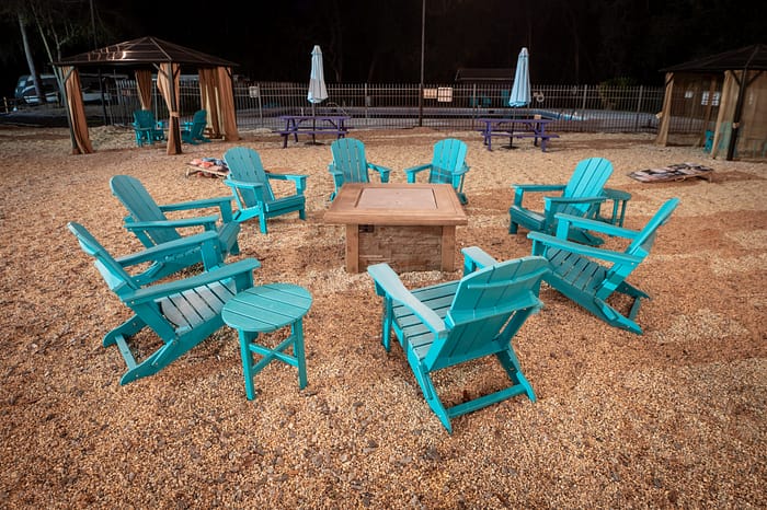 A group of turquoise Adirondack chairs arranged in a circle around a fire pit on a gravel surface, with gazebos, picnic tables, and umbrellas in the background at night.