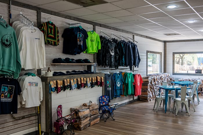 A gift shop with racks of colorful t-shirts, sweatshirts, hats, and bandanas on the left wall. On the right, there are tables and chairs by windows letting in natural light. The shop has wooden floors and a cozy atmosphere.