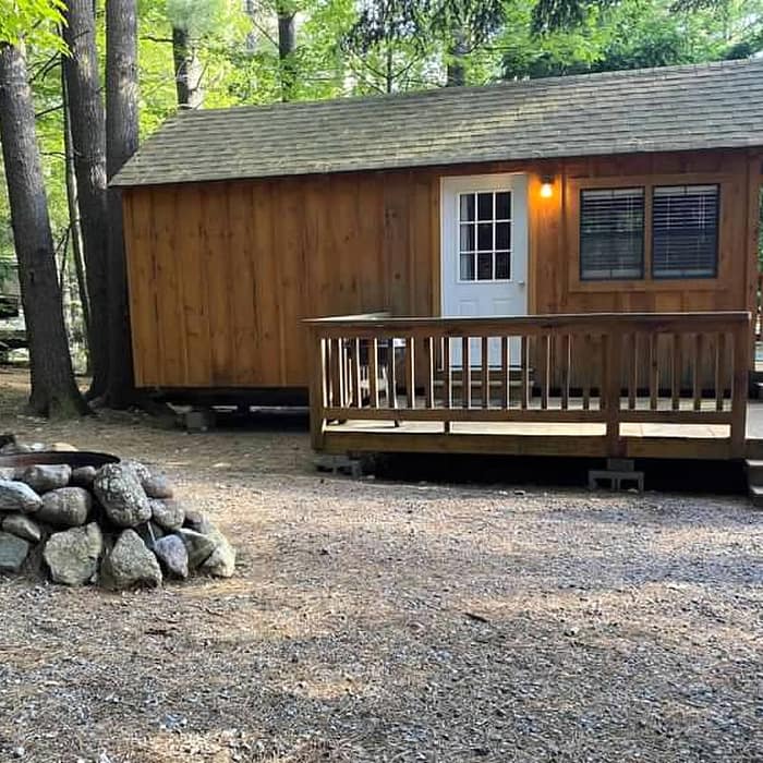 A wooden cabin with a front porch in a forested area, featuring a stone fire pit in the foreground.