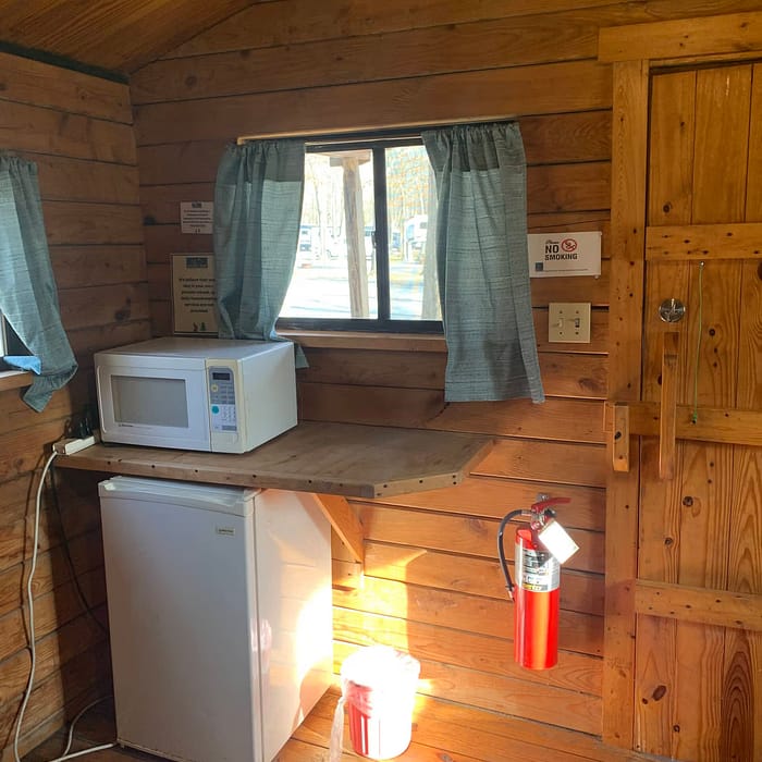 Interior of a wooden cabin with a refrigerator, microwave, small counter, window with curtains, and a fire extinguisher.